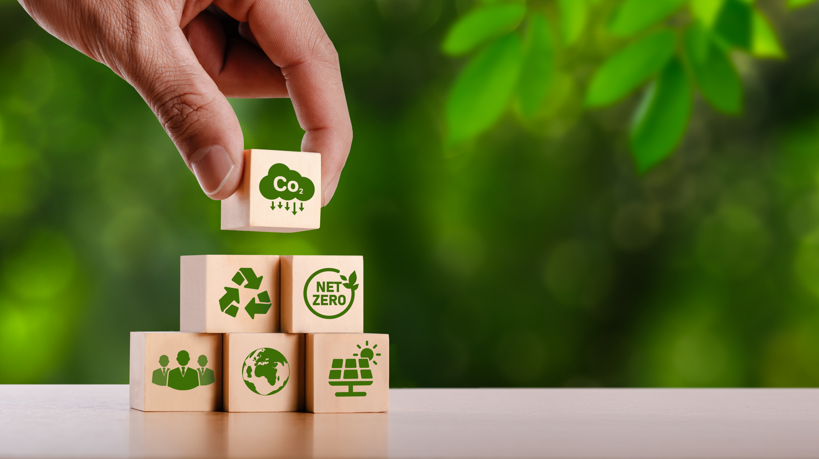 mano coloca cubos de madera uno sobre otro haciendo una pirámide. Los cubos tienen los íconos de reciclaje, figura humana, CO2, panel solar, y un planeta Tierra.